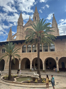 Patio in front of the facade of the Cathedral of Santa Maria of Palma with two palm trees and a central fountain featuring a lion sculpture and cacti